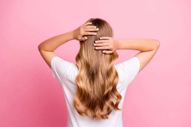 Woman dressed in a stylish t-shirt with hands touching her wavy hairdo, isolated on a pink-colored background. hair oil