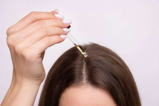 A young woman applies a drop of hair oil from a pipette to her scalp, close-up