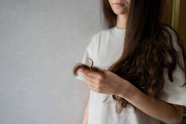 A woman appears concerned as she closely inspects the split ends of her long, wavy brunette hair oil.