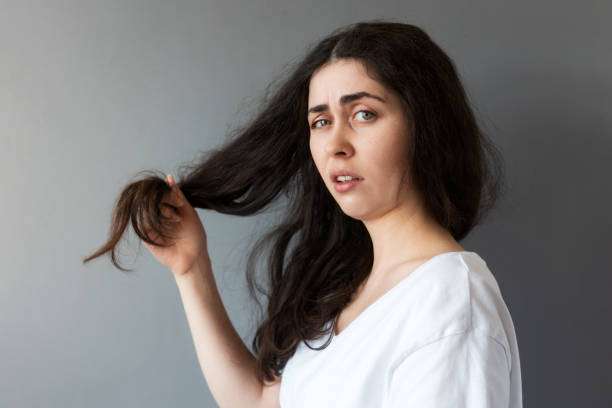 A portrait of a young upset woman shows dry tips of her long dark hair. A lady dripping hair oil with a pipette on her hair.