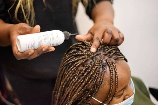 Hands of a hairdresser putting hair oil on head of an African female client with braided hair in salon during pandemic