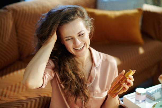 A smiling stylish housewife in pajamas with long brunette wavy hair holding a hair oil bottle in the modern house on a sunny winter day.