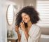 Happy young woman in bathrobe styling her hair oil in front of mirror in bathroom