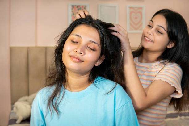 her head massaged by her female friend after applying hair oil for strong and shiny hair growth and enjoying their hair care moments in the bedroom.