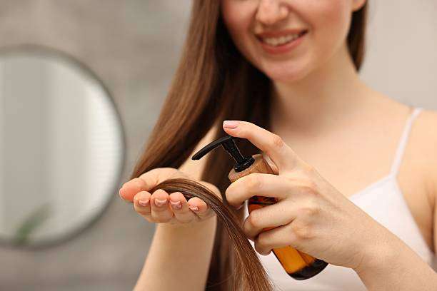 Smiling woman applying leave-in hair oil from a pump bottle to her long, straight hair.